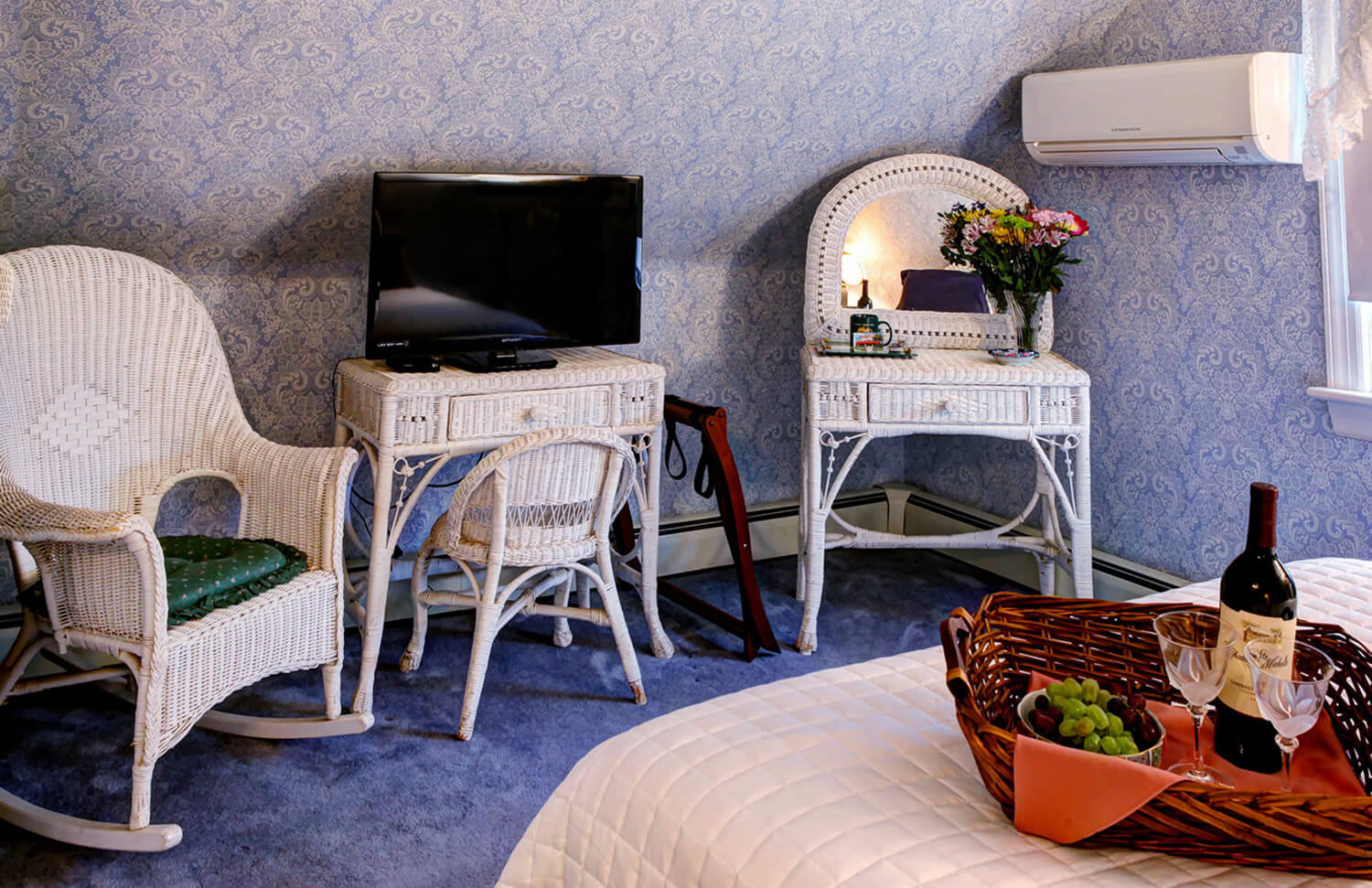 bed with white quilt and a brown basket with fruit and wine, white rocking chair and TV