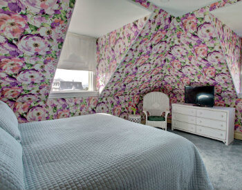 king bed with beige floral quilt and multi-colored hydrangea wallpaper, corner fireplace and TV sitting on white wicker dresser