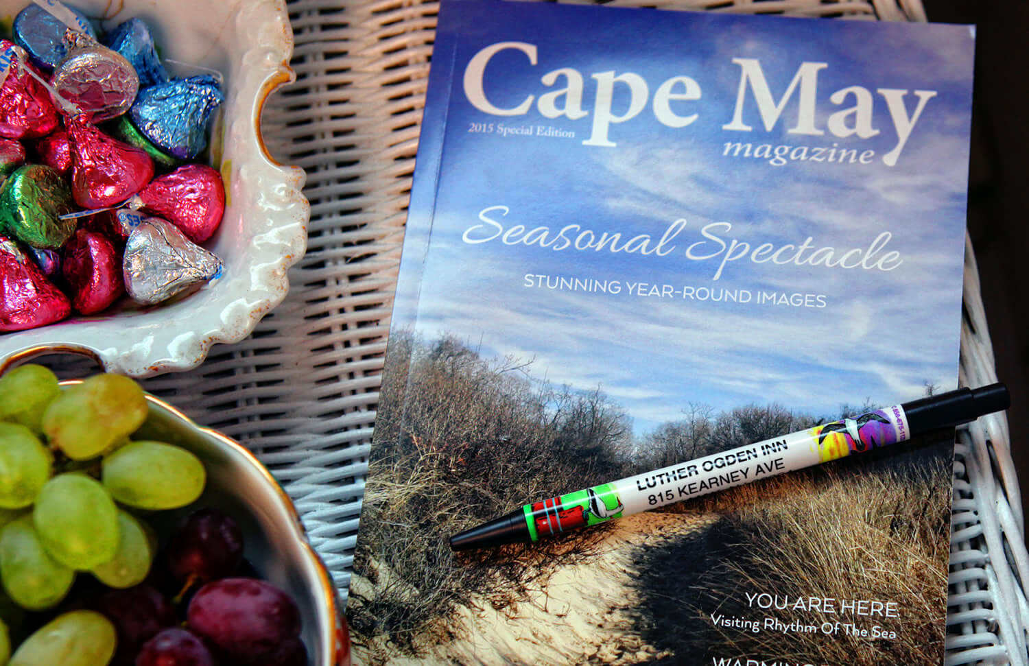 Sitting on a white wicker table is a bowl of candy, a pen, red and green grapes and a magazine