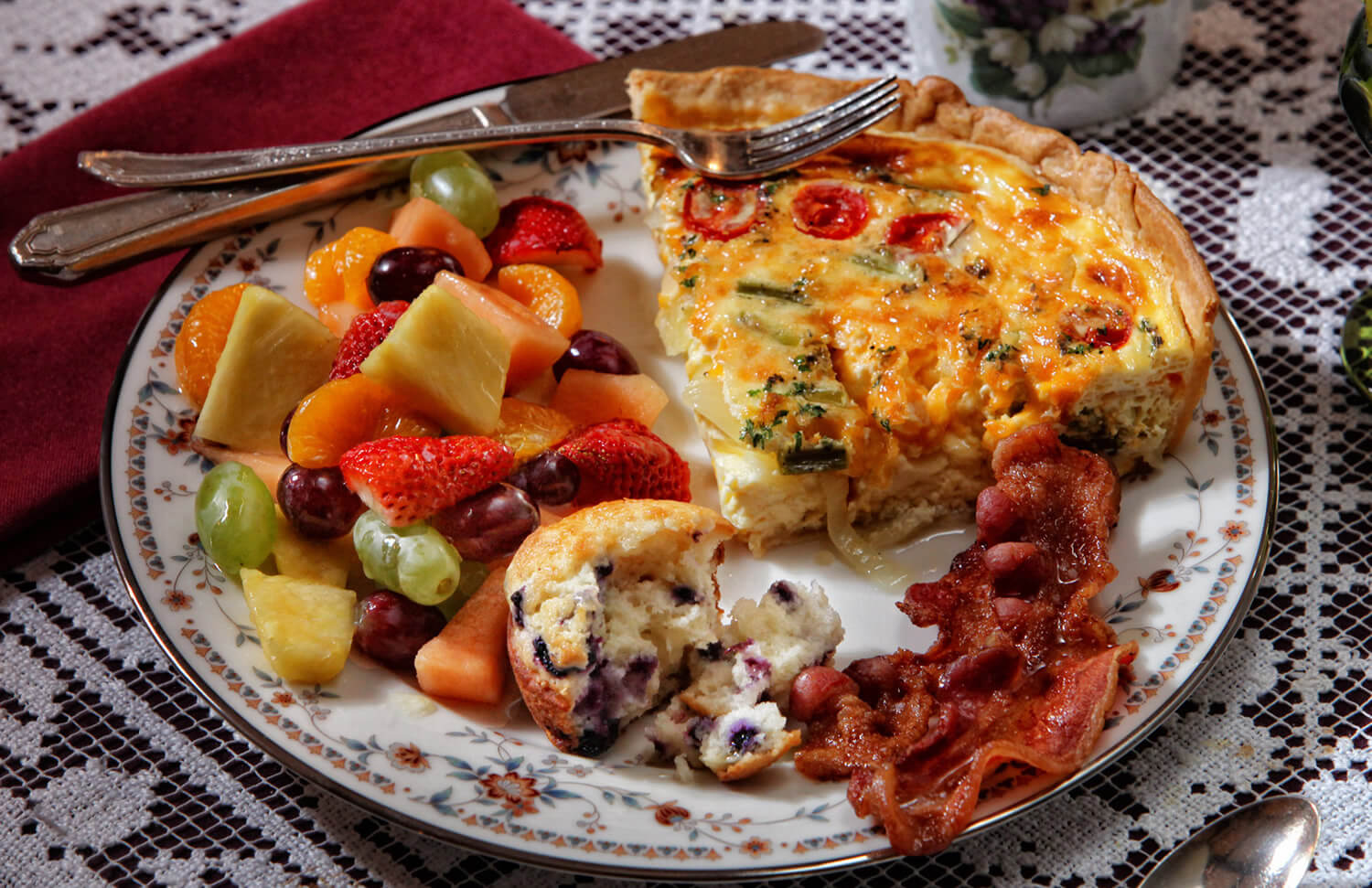 wedge of ham and cheese quiche, blueberry muffin, bacon and red strawberries, orange/ green melon, red/ green grapes