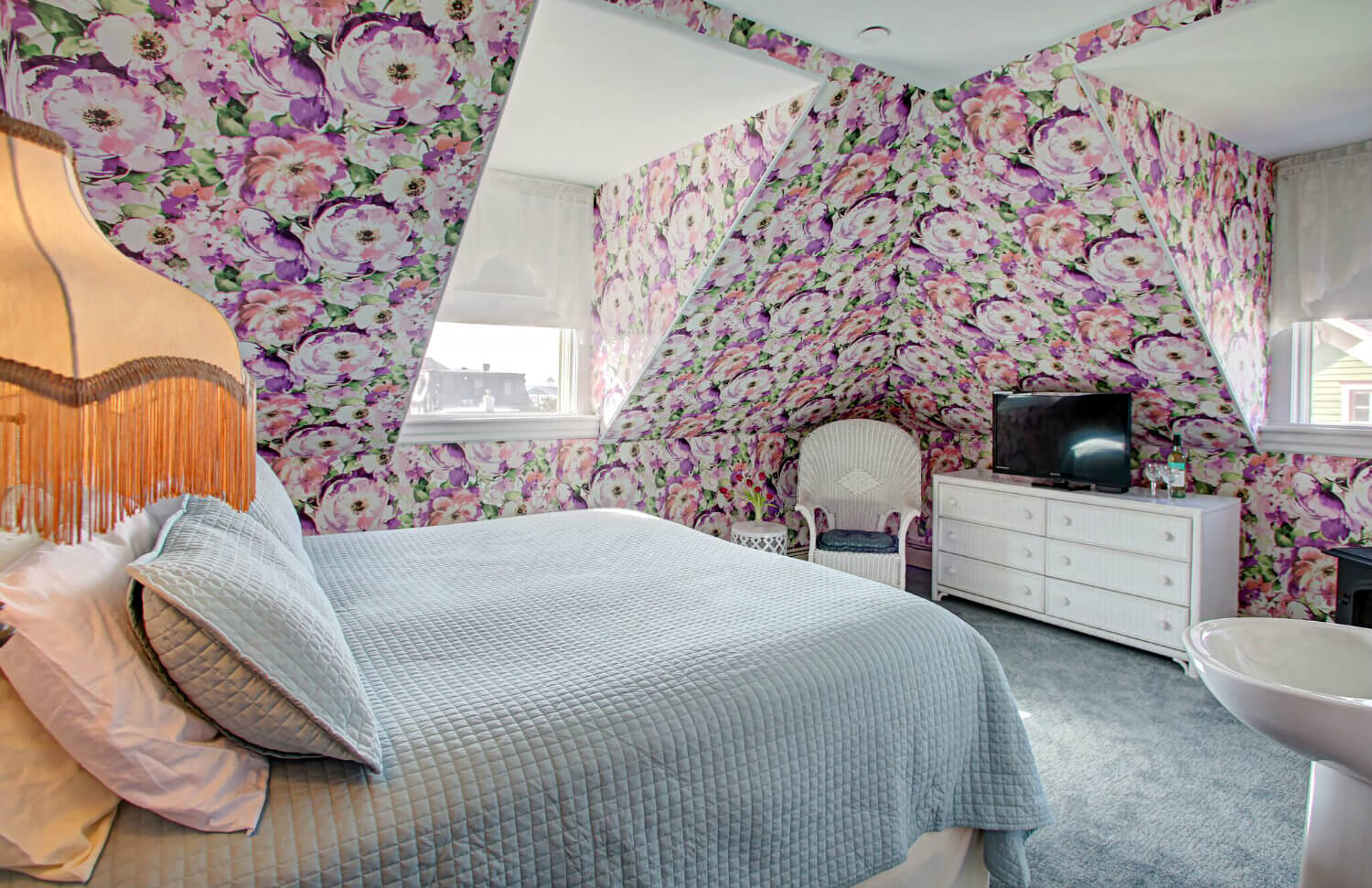 king bed with beige floral quilt and multi-colored hydrangea wallpaper, corner fireplace and TV sitting on white wicker dresser