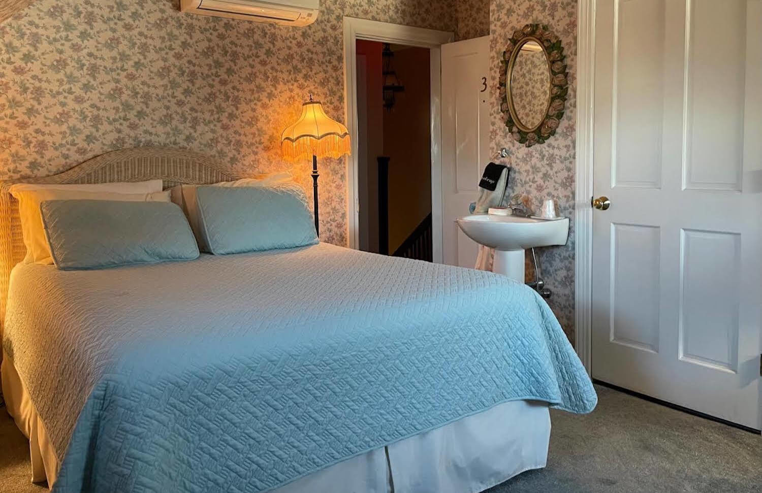Queen bed with green quilt, white pedestal sink with oval mirror above, floor lamp, flowered wallpaper, air conditioner unit above bed