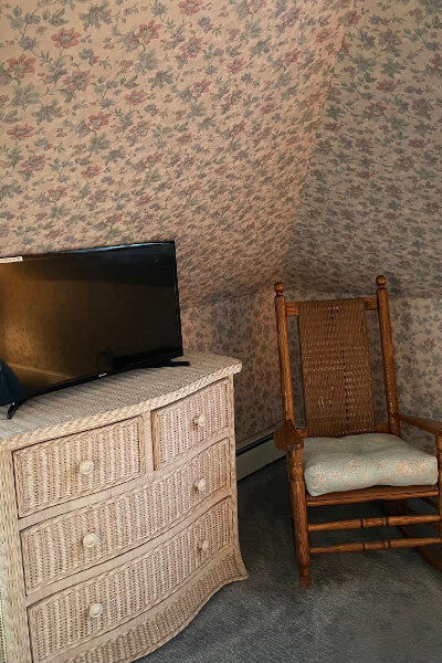 White wicker stand with drawers holding flat-screen television in room with floral wallpoaper and cushioned rocking chair in corner.