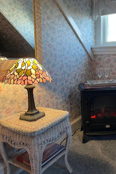 White wicker nightstand holding lamp with mirror above in room with floral wallpaper, cherry stamp holding tray with glasses under window.