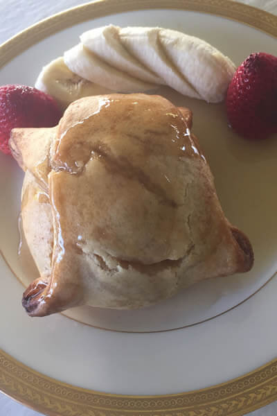 baked apple dumpling on a white plate with gold rim with sliced bananas and 2 strawberries